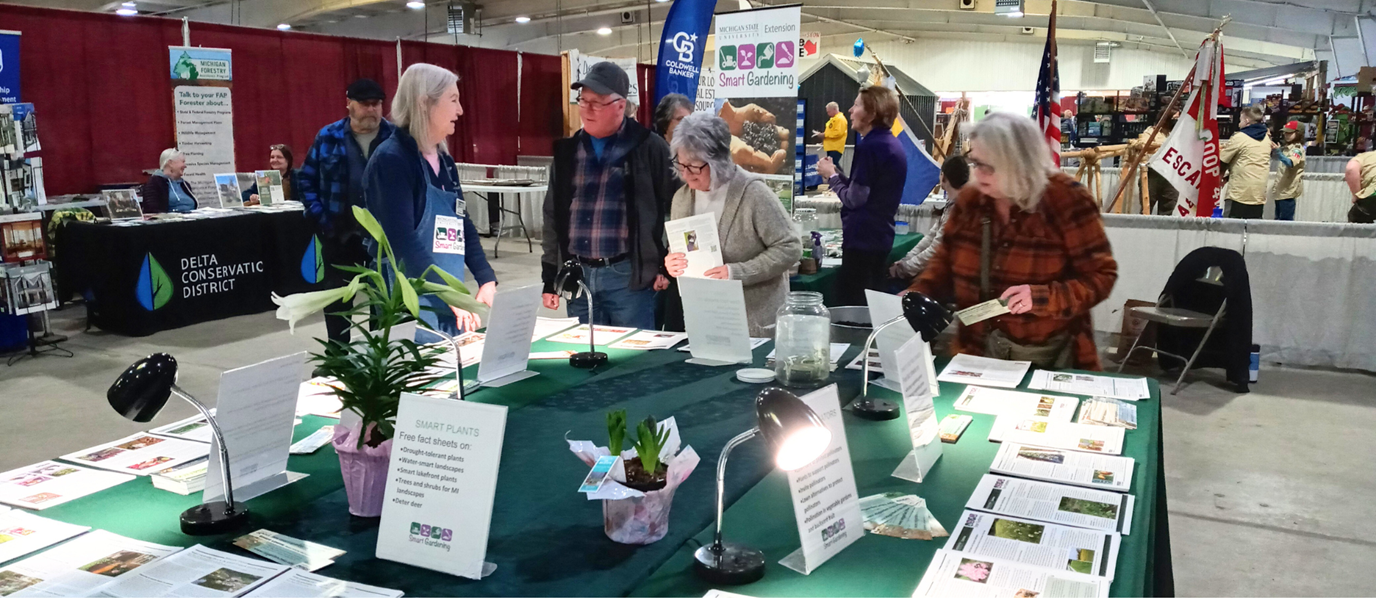 A community outreach event featuring the MSU Extension Smart Gardening initiative. Several people are gathered around a table covered with educational materials and potted plants, engaging in conversation with Master Gardeners. Signs on the table promote free fact sheets on smart plant choices, and lamps illuminate the resources. In the background, other booths such as Delta Conservation District and Michigan Forestry are visible, along with attendees browsing the exhibition space.